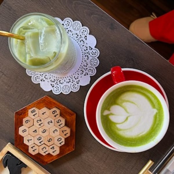 Overhead view of an iced matcha latte, a hot matcha latte with latte art, and a wooden puzzle.