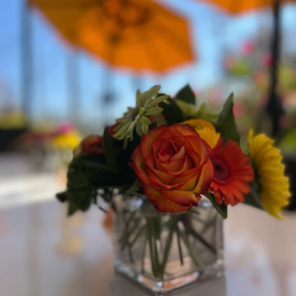 A vibrant floral centerpiece with orange and yellow flowers sits on a table, with orange umbrellas in the background.