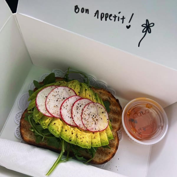 Avocado toast with radishes and arugula in a takeout box with a side of sauce.
