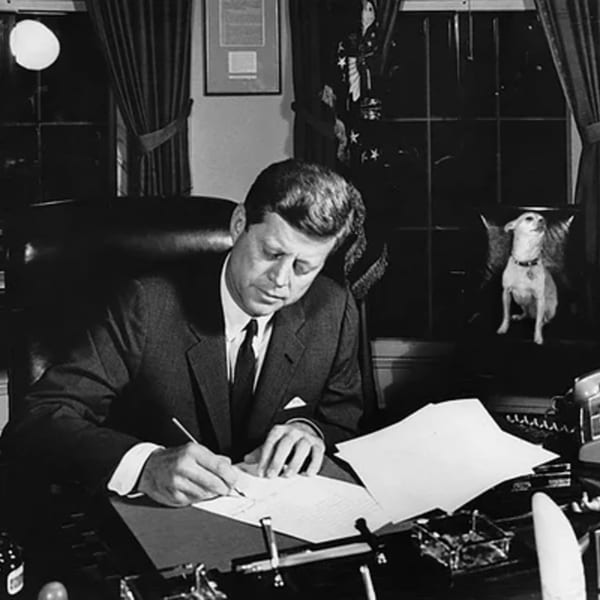 A man in a suit writes at a desk, with a small dog sitting on a chair beside him.