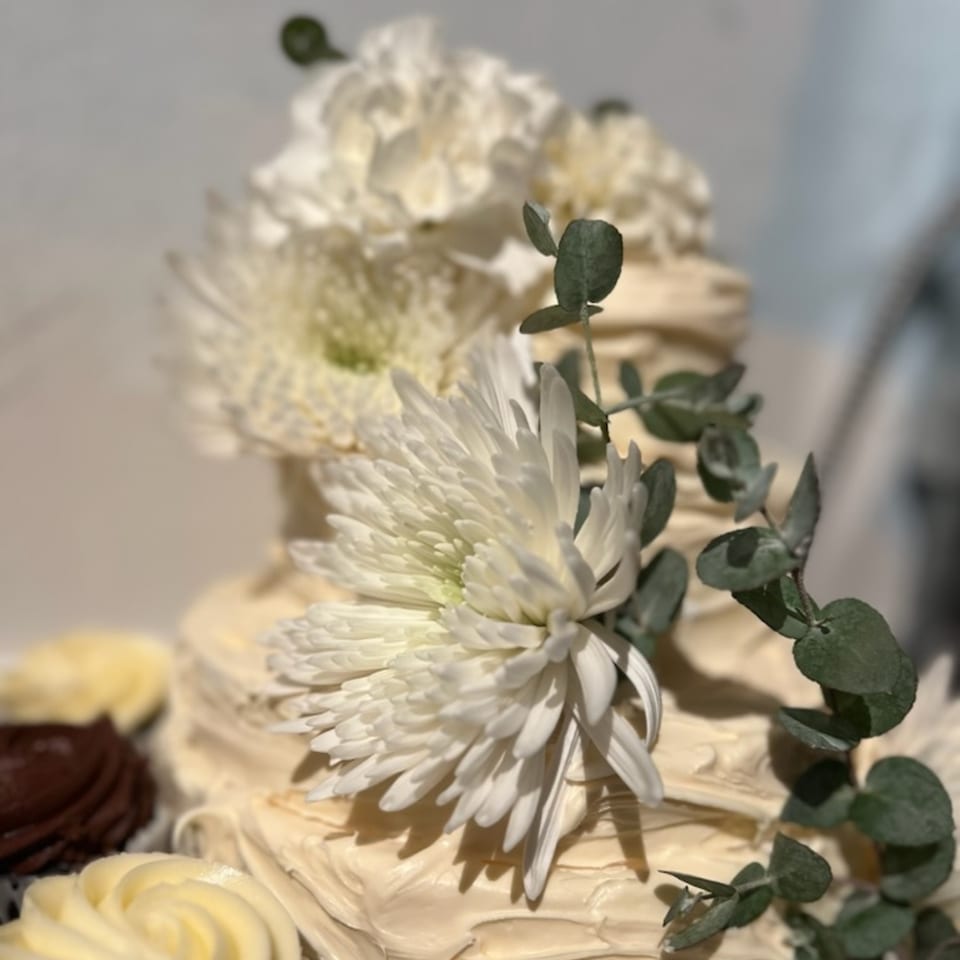Tiered cake with white frosting, decorated with white flowers and eucalyptus leaves, alongside cupcakes.