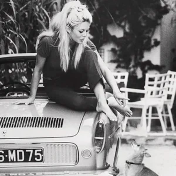 Black and white photo of a woman sitting on a car hood, looking at a dog.