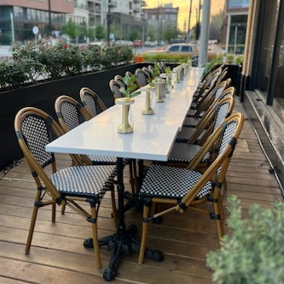 Long outdoor dining table with checkered chairs and small gold vases on a wooden deck.