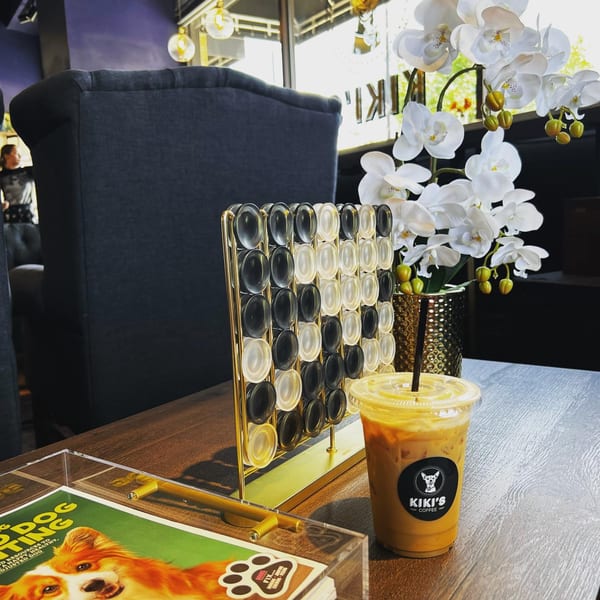 Iced coffee on a wooden table next to a Connect Four game, orchids, and a book.