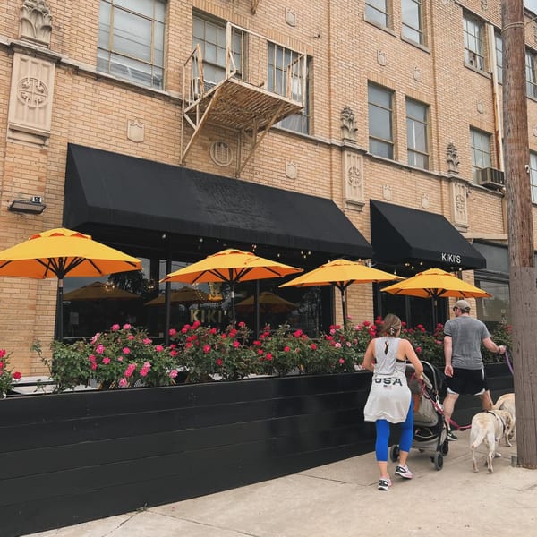 Kiki's Coffee & Spirits exterior with orange umbrellas, rose planters, and people walking dogs.