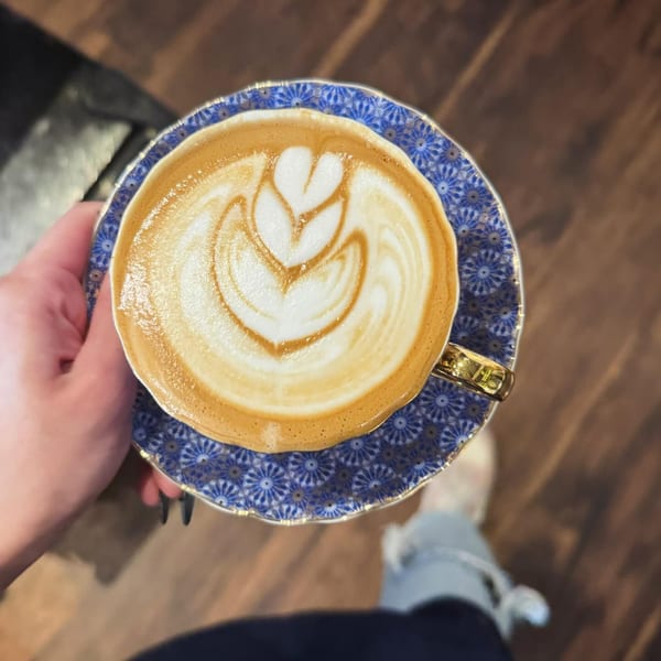 A hand holds a blue and white patterned cup of Kiki's latte with heart-shaped foam art.