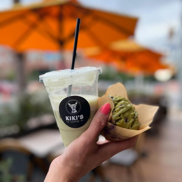 Hand holding a Kiki's Coffee iced drink and a green muffin with orange umbrellas in the background.