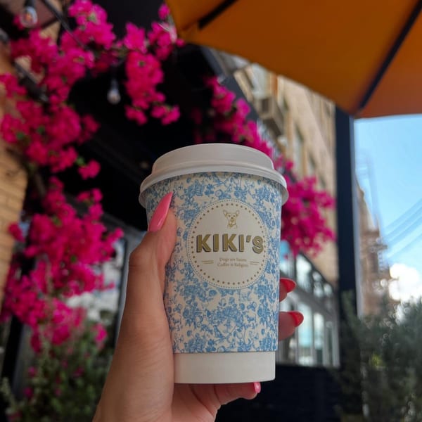 Hand holding a Kiki's coffee cup with pink bougainvillea flowers and an orange umbrella in the background.